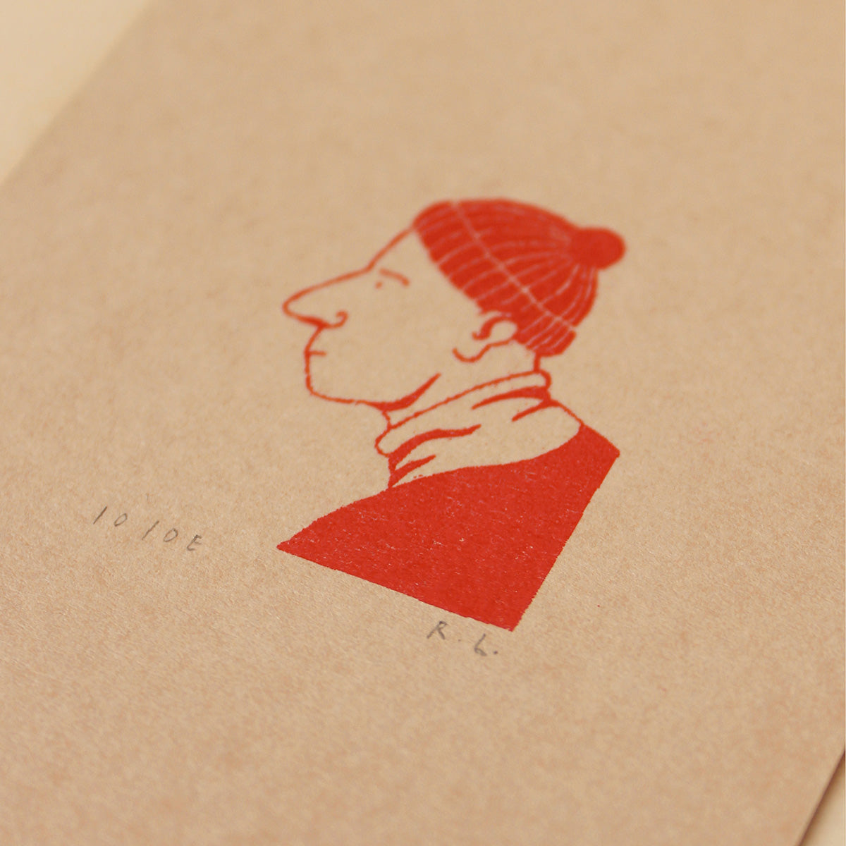 A close-up of a textured beige paper surface featuring a red ink stamp illustration of a person in profile wearing a knit beanie, with small handwritten markings nearby.