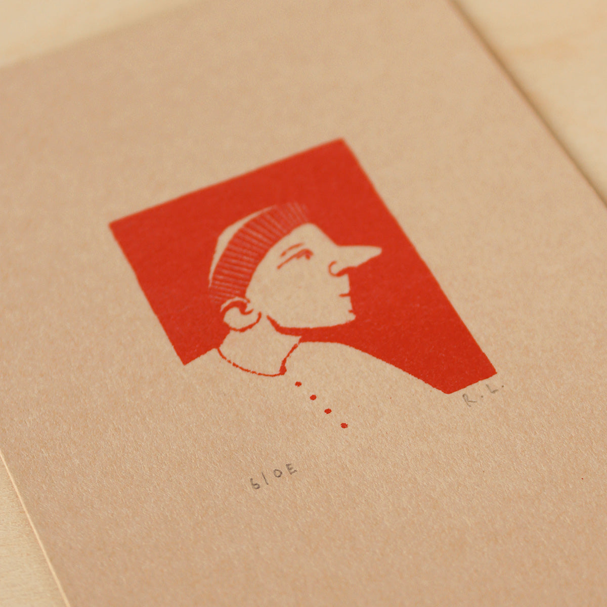 A close-up of a textured beige paper card featuring a minimalist red graphic print of a portrait of a beret wearing man, with small printed text below.