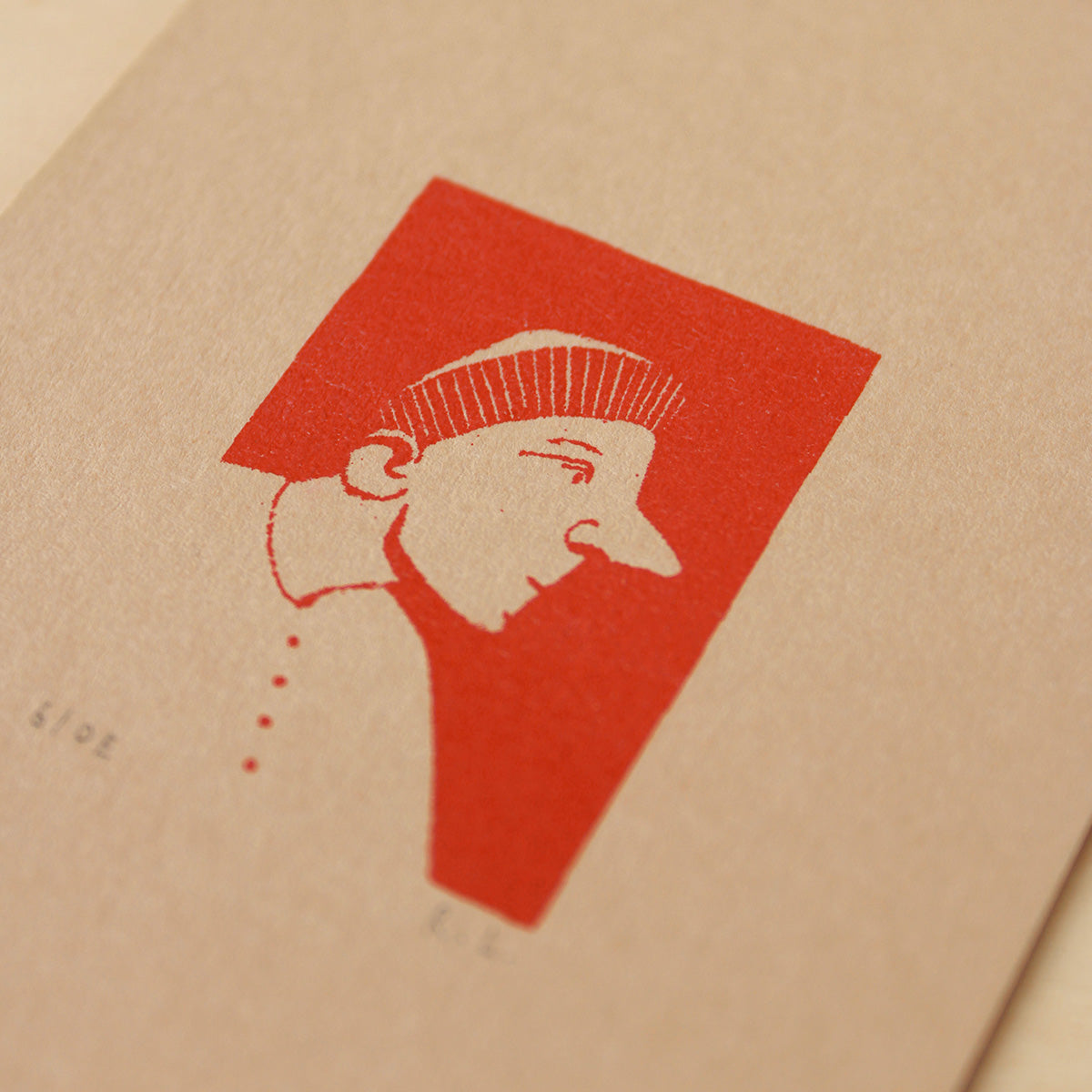 A close-up of a textured kraft paper surface featuring a minimalist red graphic print of a stylized face profile with a beret hat.