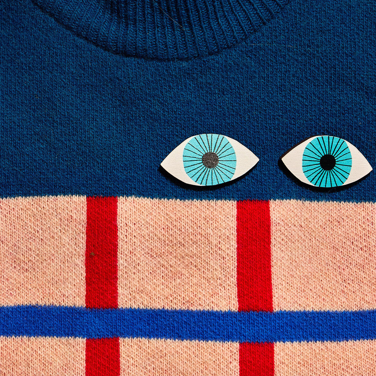 Close-up of a blue sweater with cartoon eye pin badges and red and beige stripes below.
