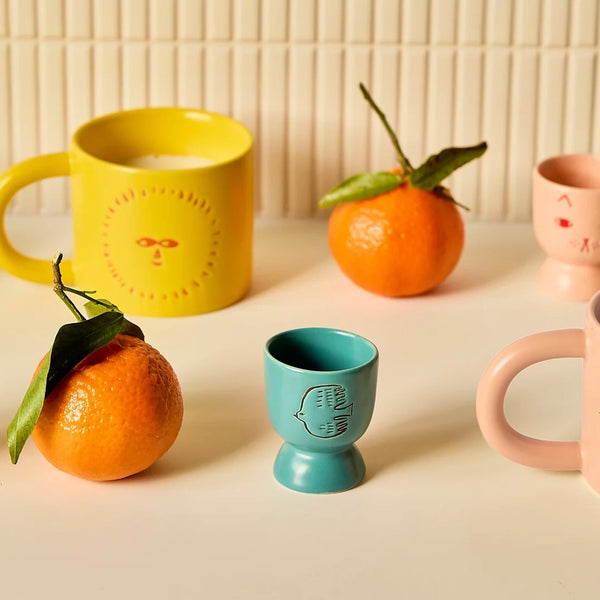 Colourful yellow, blue and pink ceramic mugs and egg cups with face, bird, sun and cat designs on a light surface with oranges.
