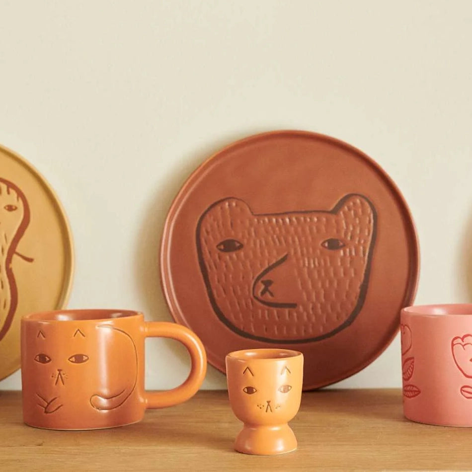 A set of handcrafted ceramic tableware in terracotta and peach tones, featuring embossed animal faces including a bear, a dog, a cat, and a duck, displayed on a wooden surface against a cream background.