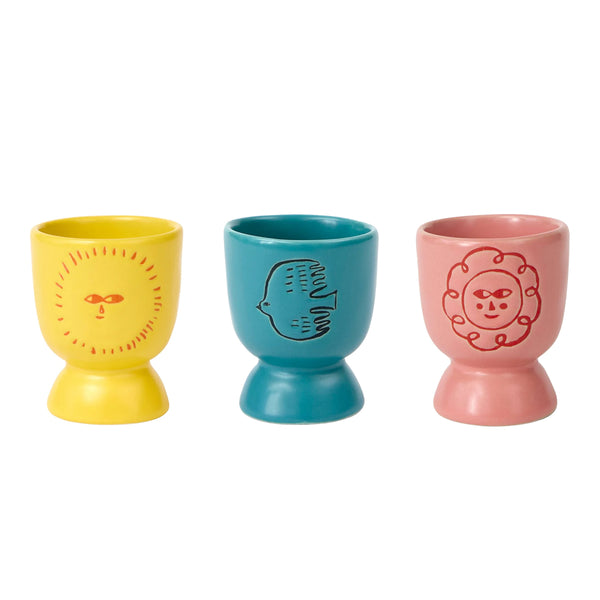 Three ceramic egg cups in yellow, teal, and pink, each decorated with a stylized face motif, shown on a plain white background.