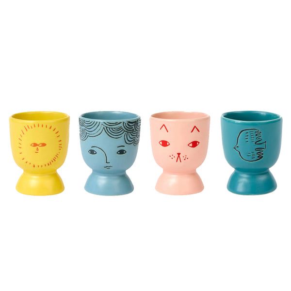 Four colourful yellow, blue, pink and teal ceramic cups with face, sun, cat and bird designs on a white background