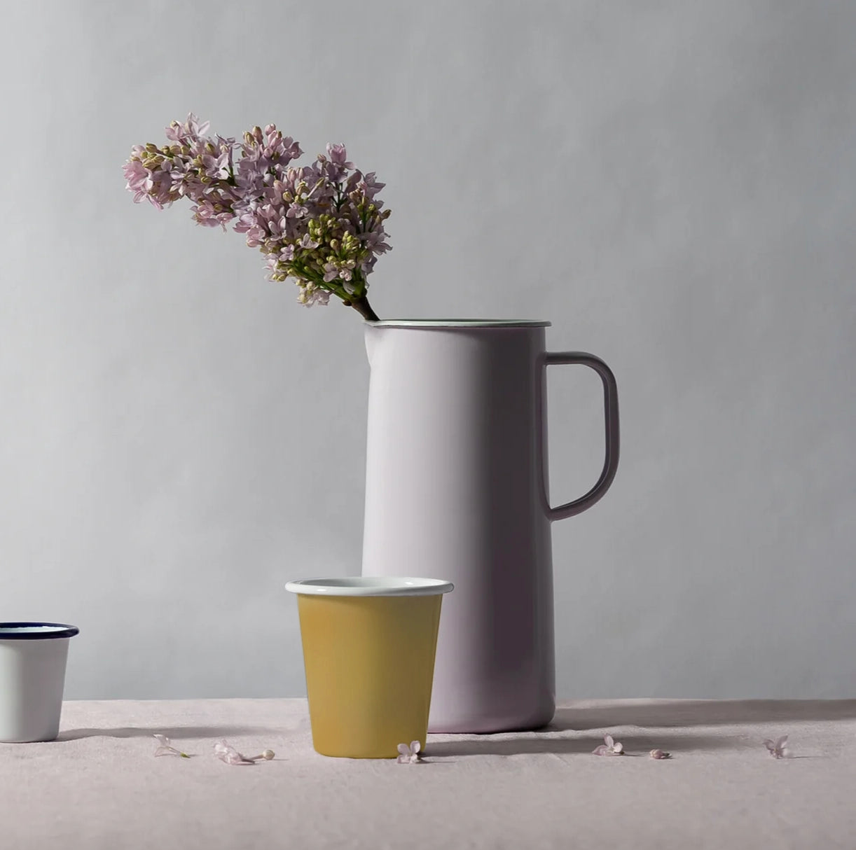 Mustard yellow falconware enamel tumbler with white rim on a table sitting with a pale lilac enamel jug with a flower in.
