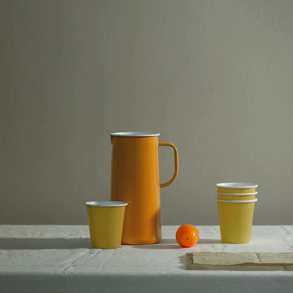 Orange tangerine falconware enamel jug  on a table setting with linen tablecloth and mustard yellow enamel tumblers. 