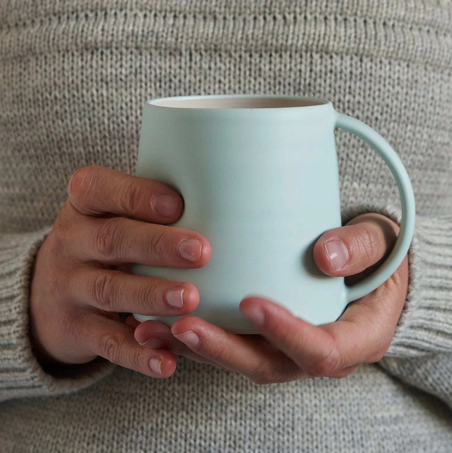 Person holding a light blue mug against a grey jumper.