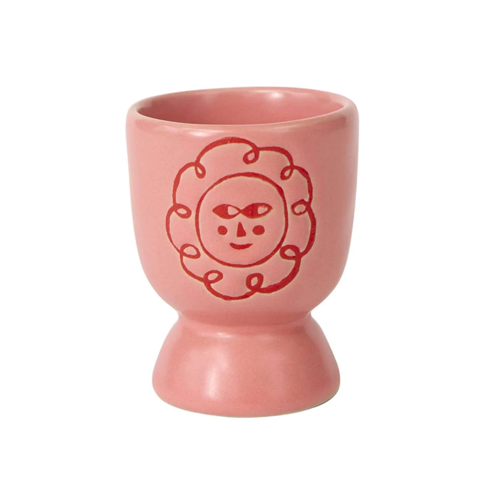 A pink ceramic egg cup with a red line-flower face design on the front, set against a plain white background.