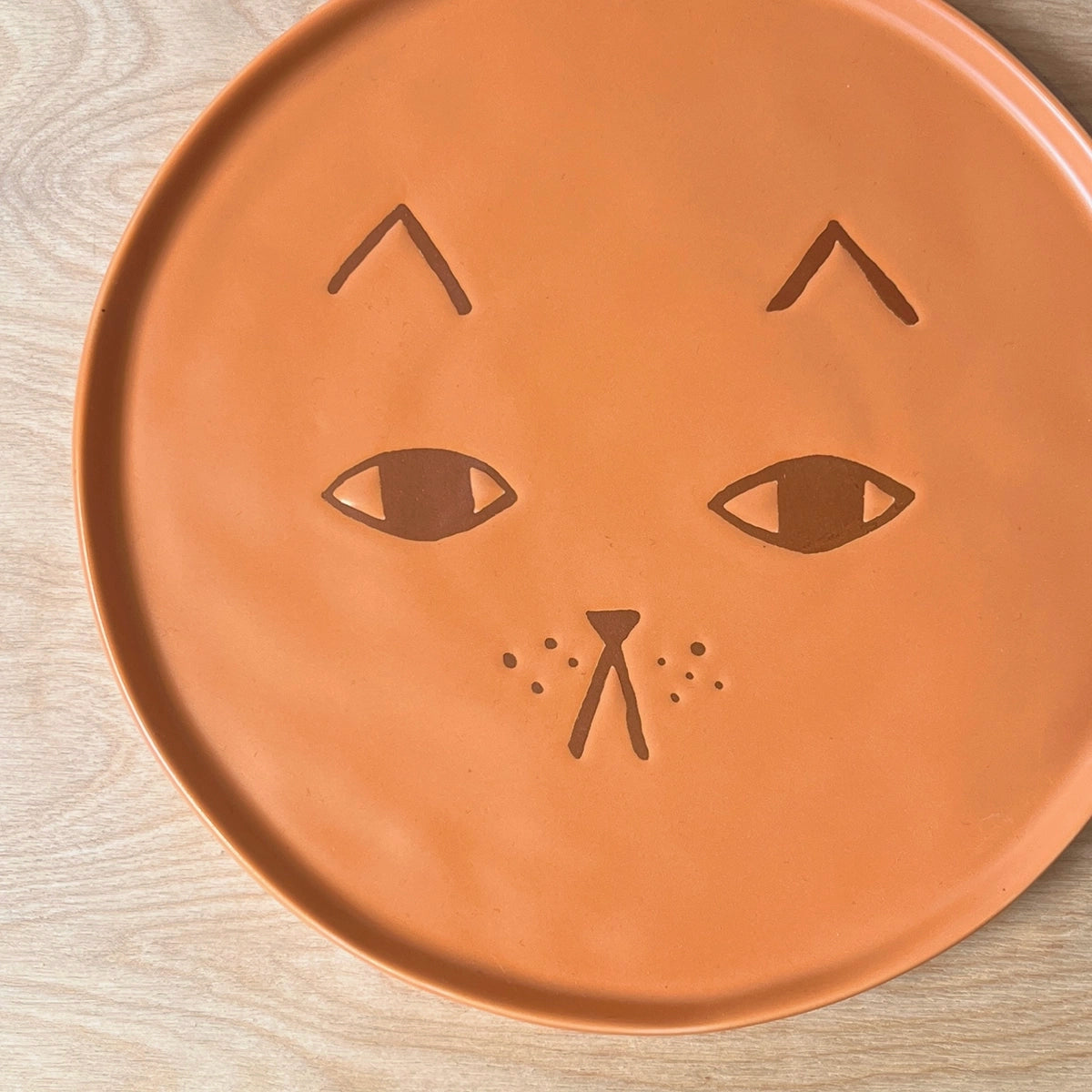 A round terracotta-colored ceramic plate featuring a carved cat face design, shown from a top-down view on a light wood grain surface.
