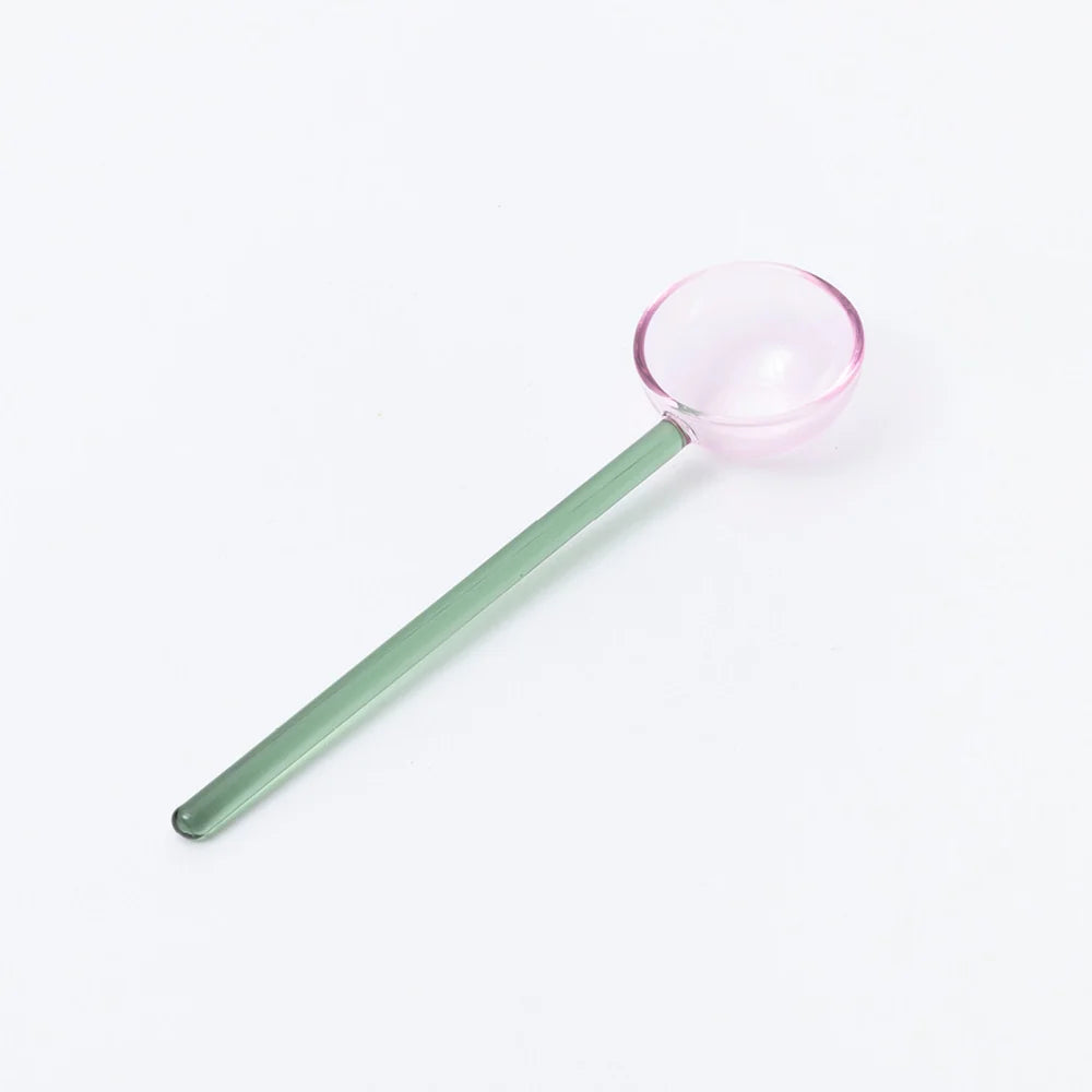 A translucent glass spoon with a long sage green handle and a round pink-tinted scoop, shown diagonally on a clean white background.