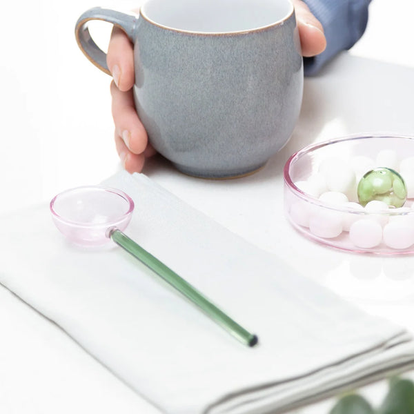 A long-handled glass spoon on a white napkin, with a translucent pink scoop and green handle, on a white table with a coffee cup and bowl of mints.