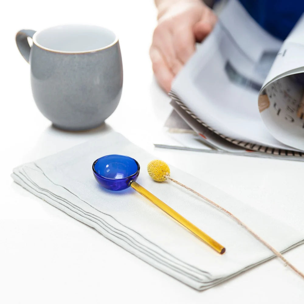 A long-handled glass spoon on a white napkin, with a translucent blue scoop and yellow handle, on a white table with a coffee cup and someone reading a magazine.