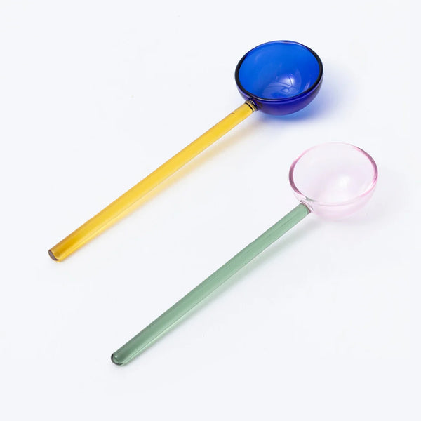 Two long-handled glass spoons on a white background, one with a translucent blue scoop and yellow handle, and one with a translucent pink scoop and green handle.