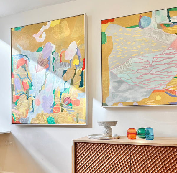 A gallery interior with two large abstract paintings in mustard colours, with a wooden cabinet hosting a range of glassware and ceramics