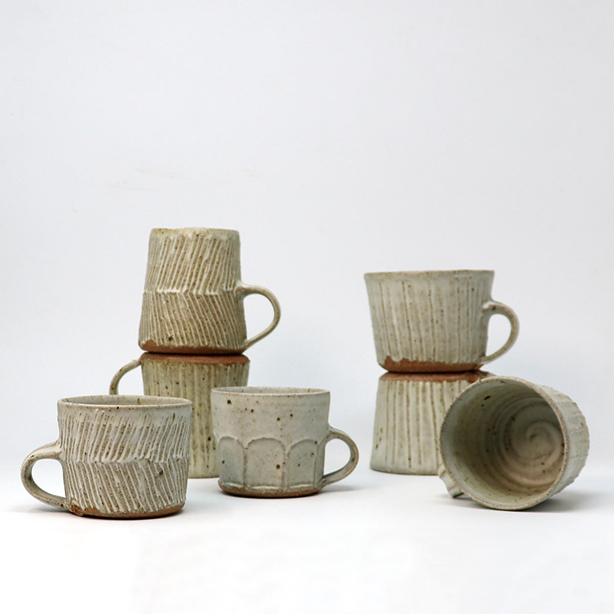 A group of handmade ceramic mugs in a light beige and brown glaze, arranged on a white studio background. Some mugs are stacked, and one is shown lying on its side to display the interior texture.