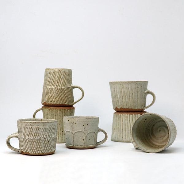 A group of handmade ceramic mugs in a light beige and brown glaze, arranged on a white studio background. Some mugs are stacked, and one is shown lying on its side to display the interior texture.