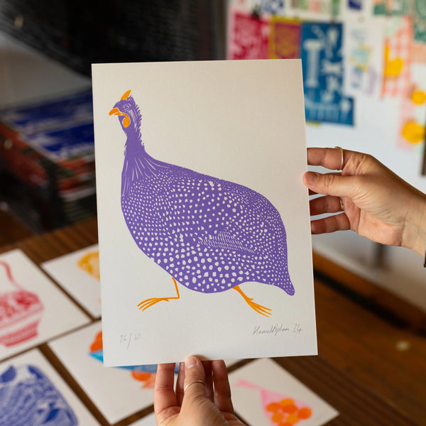 A person holds up a white art print featuring a purple and orange spotted bird illustration, with other colorful prints visible on a wooden table in the background.