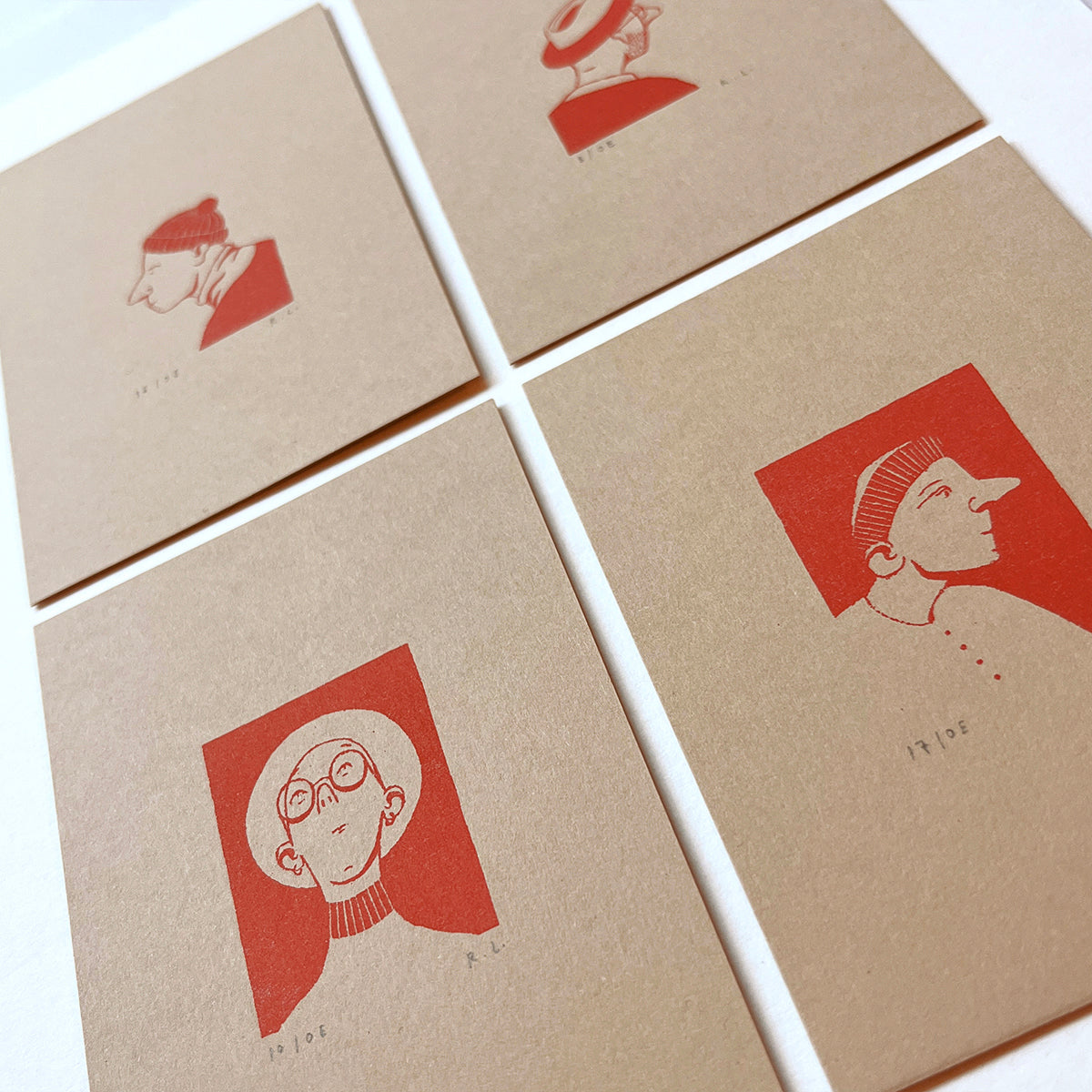 Four square kraft paper cards arranged in a grid on a white surface, each printed with a minimalist red illustration of a portraits of hat wearing people and small text.