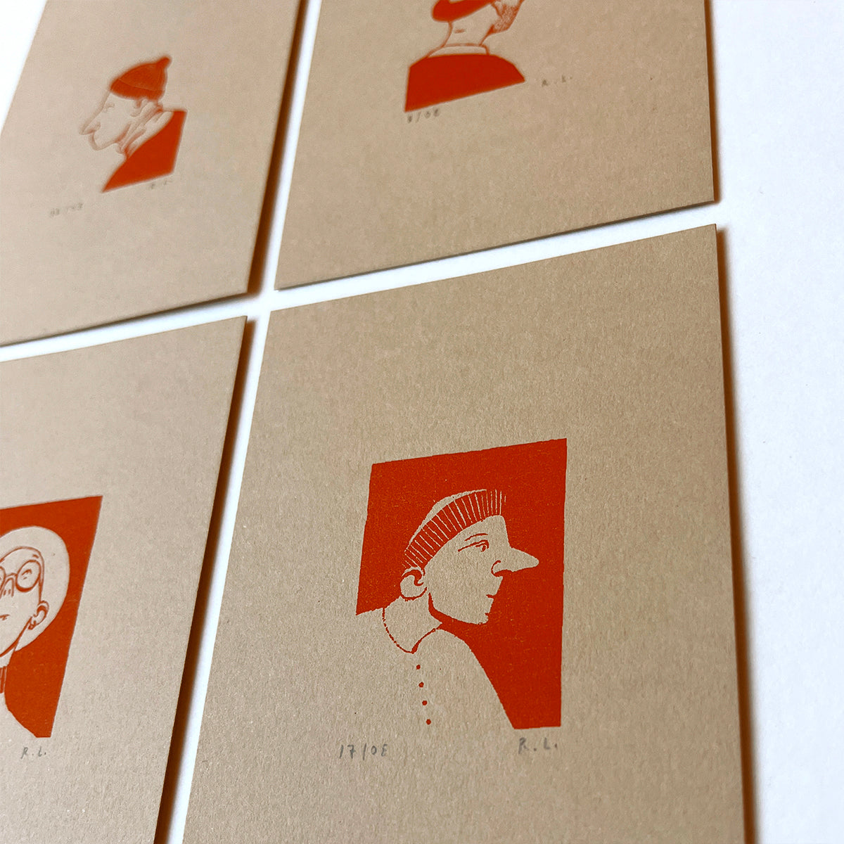 A close-up of several square kraft-paper cards arranged in a grid on a white surface. Each card features a minimalist orange-red profile hat portrait illustration with small handwritten initials below.