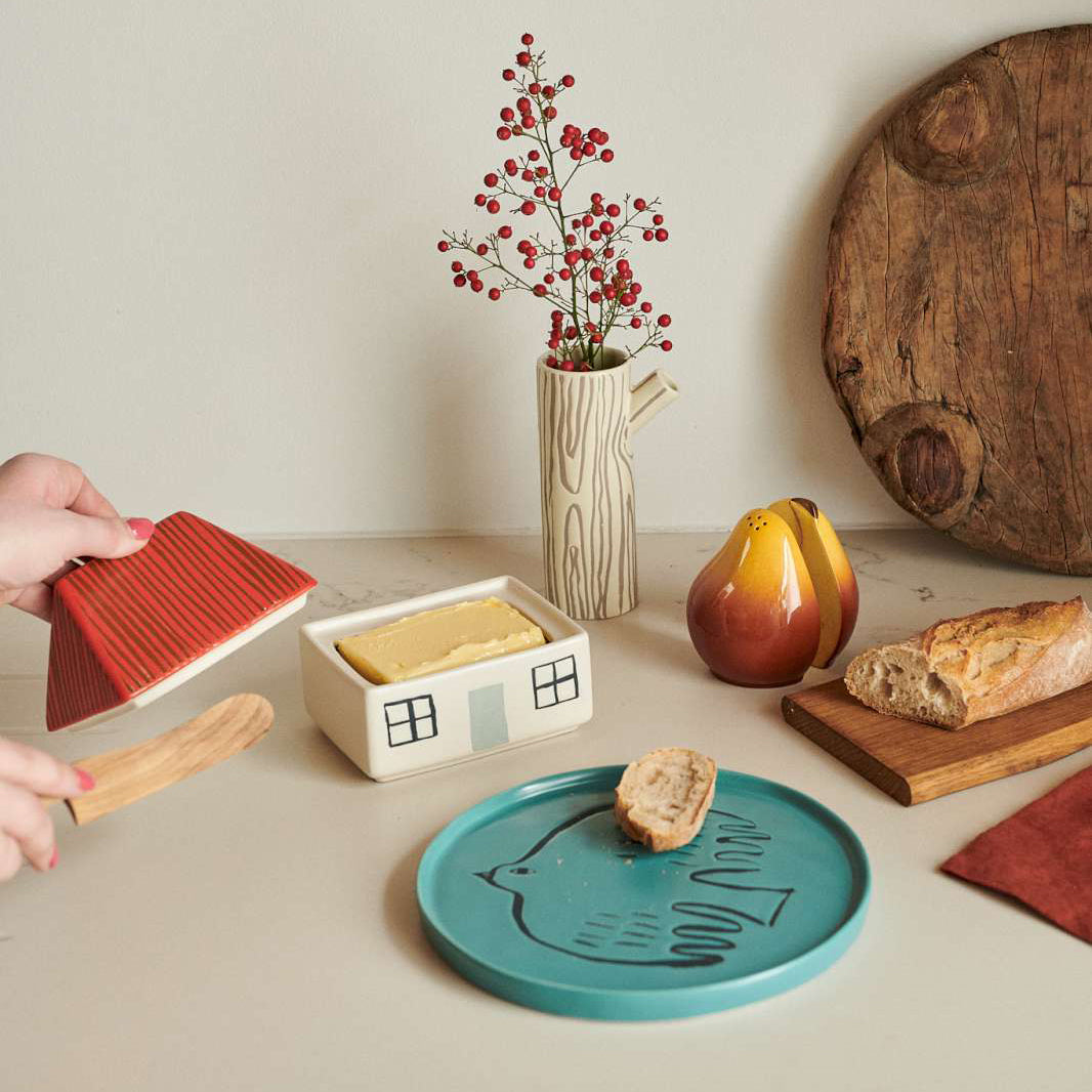 A styled tabletop scene with a hand holding a red striped ceramic butter dish lid next to a house-shaped butter dish containing yellow butter. The scene includes a turquoise plate with a bread slice, a pear, a loaf of bread on a wooden board, a vase with red berries, and a large round wooden cutting board against a beige wall.