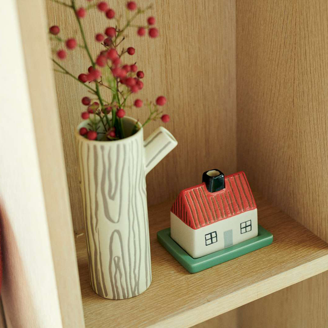 A ceramic tree stump vase with a wood grain pattern holding pink berries, displayed on a wooden shelf next to a small house-shaped ceramic container.