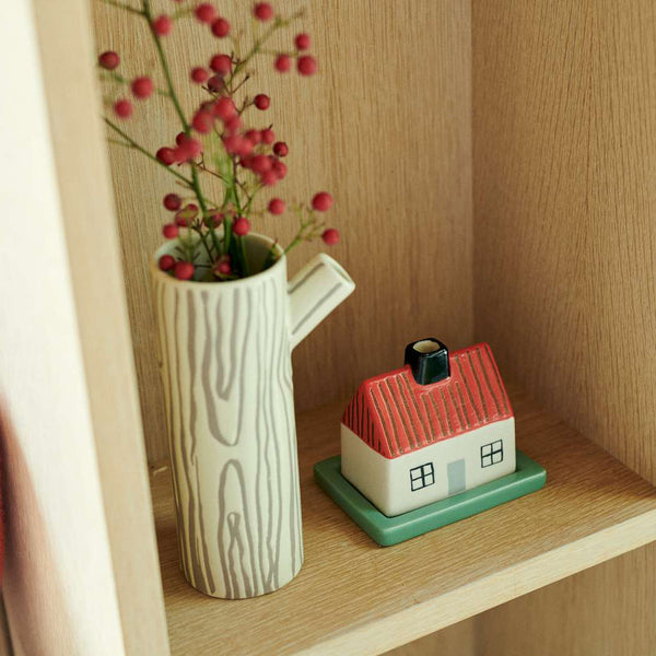 A ceramic tree stump vase with a wood grain pattern holding pink berries, displayed on a wooden shelf next to a small house-shaped ceramic container.