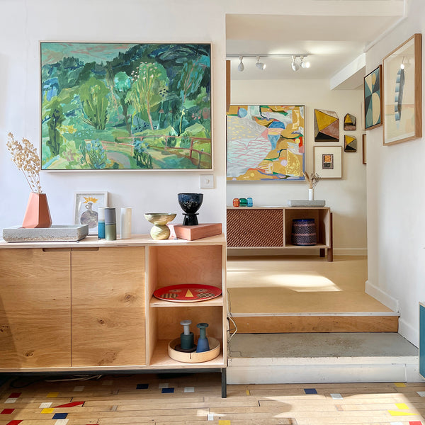 Modern interior with wooden furniture, colorful art pieces, and decorative items. Colorful landscape painting in greens with trees and a pathway painting of Costa Rica. Acrylic on canvas, framed in a white wash tray frame by Ginny Elston