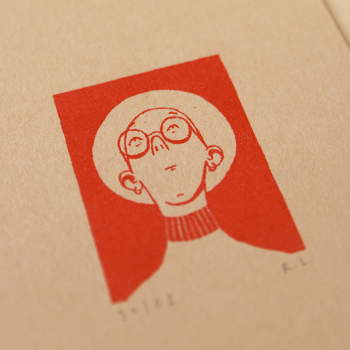 A close-up of a textured beige paper print featuring a minimalist red graphic of a person with glasses and a panama hat inside a red square, with the signature.