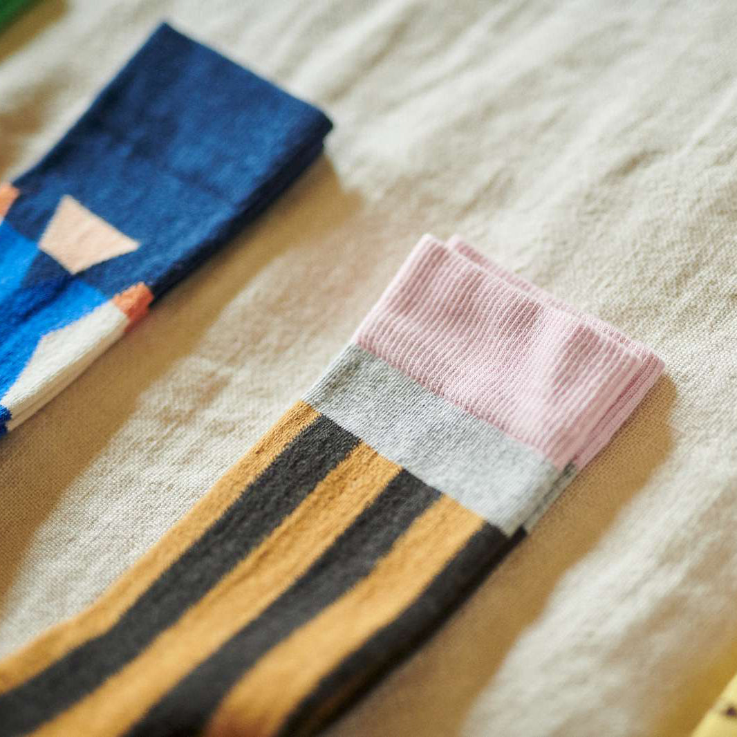 A close-up of a striped sock with a pink ribbed cuff, a light gray band, and alternating black and mustard yellow stripes, laid on a beige textured fabric surface. Another blue patterned sock is partially visible in the background.