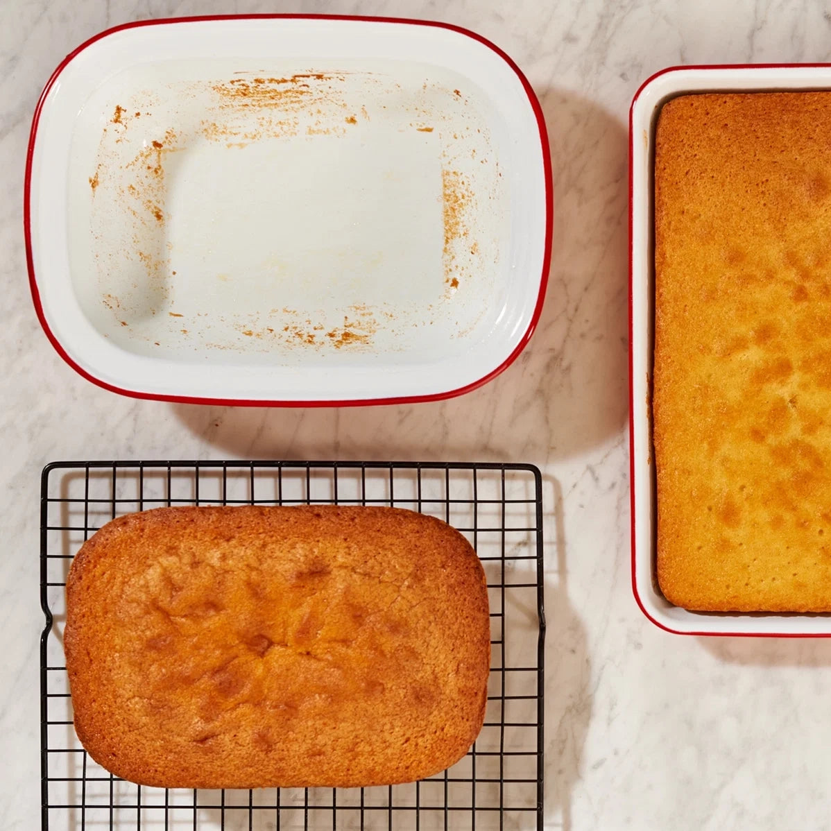 Two rectangular golden-brown baked cakes or loaves are shown on a light marble surface: one cooling on a black wire rack and one inside a white baking dish with a red rim. A second empty white baking dish with a red rim sits above.