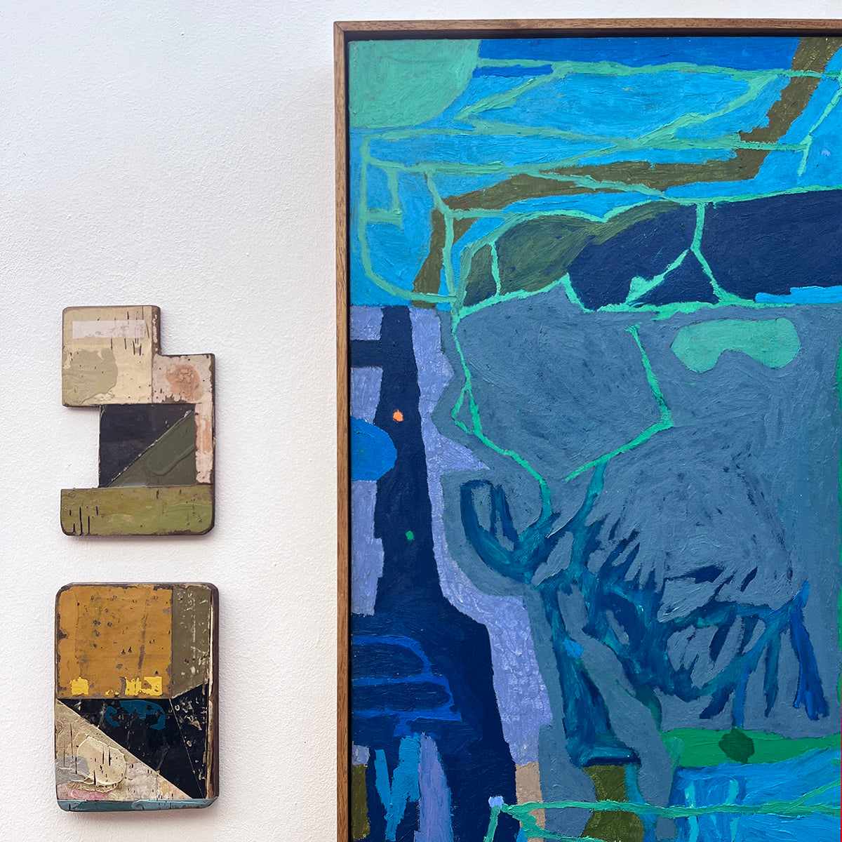 An abstract framed painting with blue, teal, and gray brushstrokes hangs on a white wall beside two small stacked abstract wooden wall pieces.
