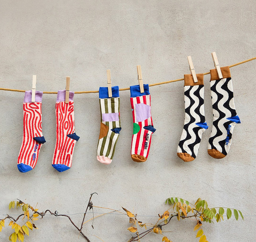 Three pairs of patterned socks hanging from a wooden clothesline with wooden clothespins against a textured light gray wall, with small branches and yellow leaves at the bottom.