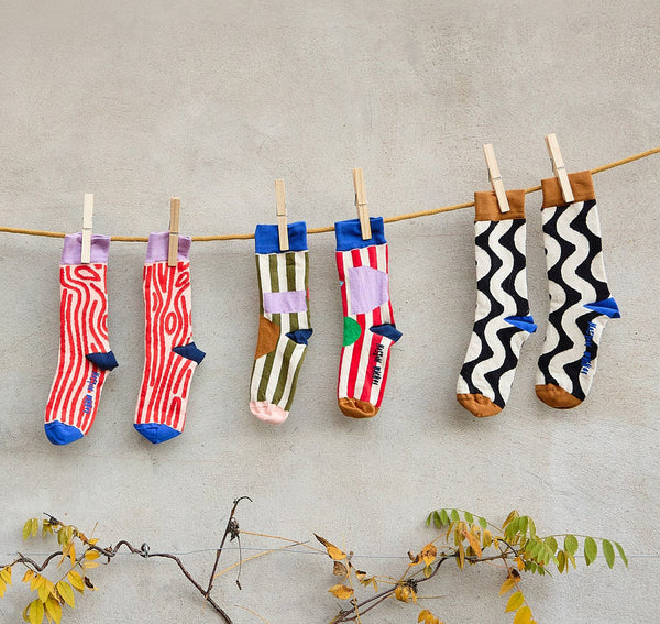 Three pairs of patterned socks hanging from a wooden clothesline with wooden clothespins against a textured light gray wall, with small branches and yellow leaves at the bottom.