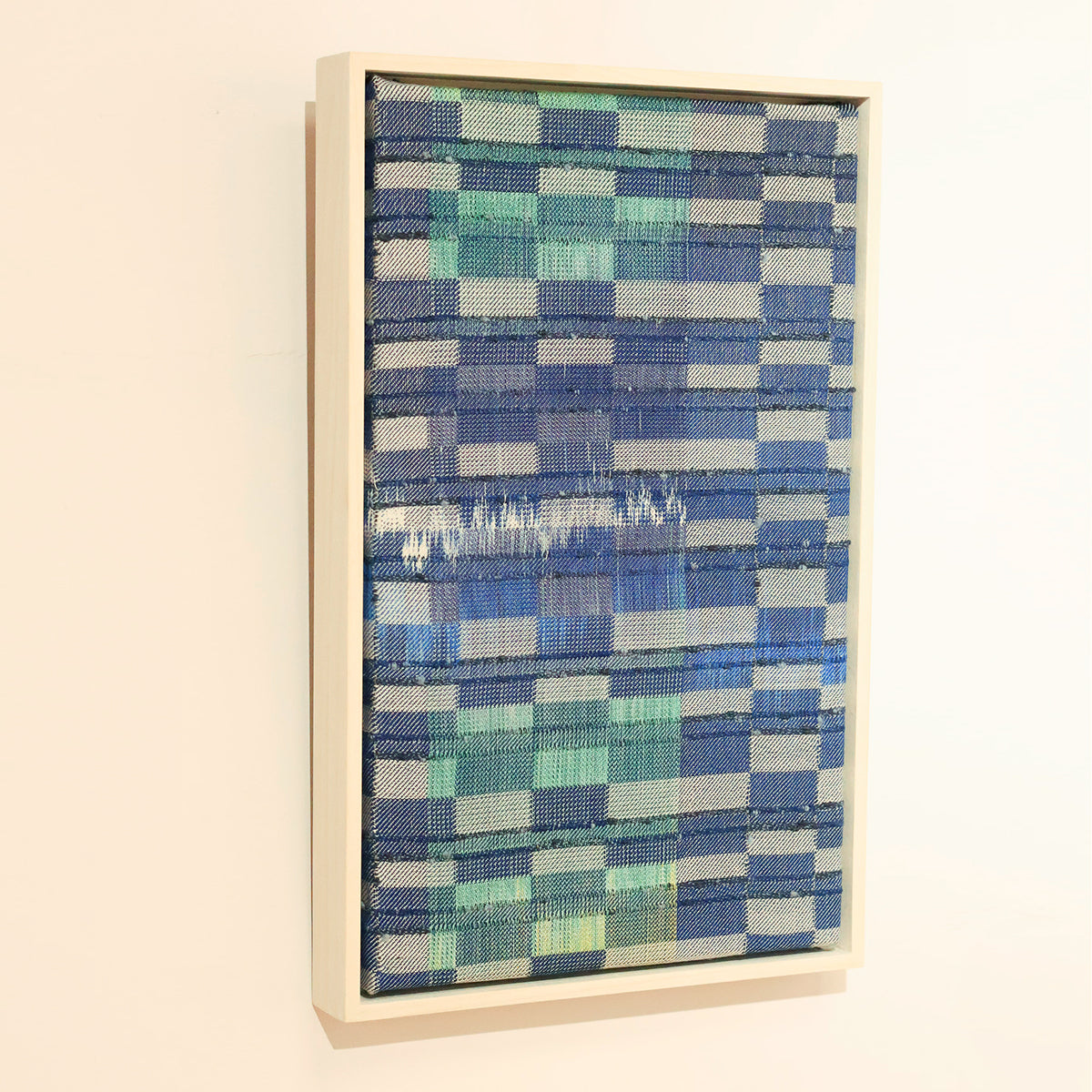 Woven Textile art piece in blue and green tones, framed in a light wooden box frame. Hung on a white wall. 