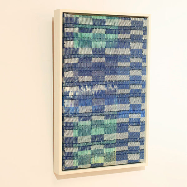 Woven Textile art piece in blue and green tones, framed in a light wooden box frame. Hung on a white wall. 