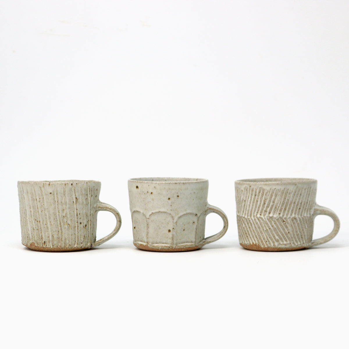 Three small handmade ceramic mugs with textured glazes in a speckled beige and off-white finish, arranged side by side on a white background.