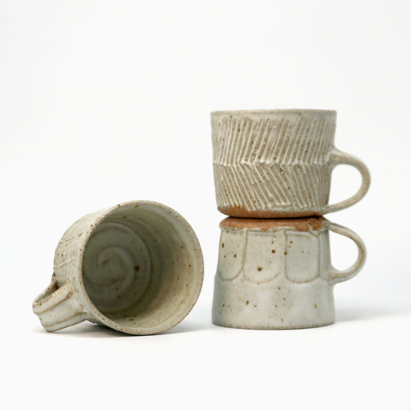 Three handmade ceramic mugs with a speckled off-white glaze and earthy brown accents, displayed on a clean white background. One mug is shown lying on its side to reveal the interior spiral design, while the other two are stacked and standing upright to show the textured exterior patterns and handles.