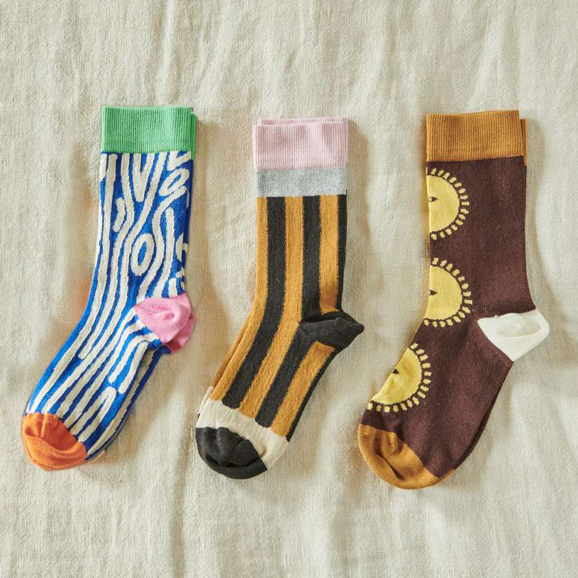 Three pairs of patterned crew socks laid flat on a light beige fabric background. The left sock has blue and white wavy stripes with a green cuff and pink heel; the middle sock has black and mustard yellow vertical stripes with a pink and light gray cuff to look like a pencil; the right sock is dark brown with yellow sun motifs and a mustard yellow cuff.