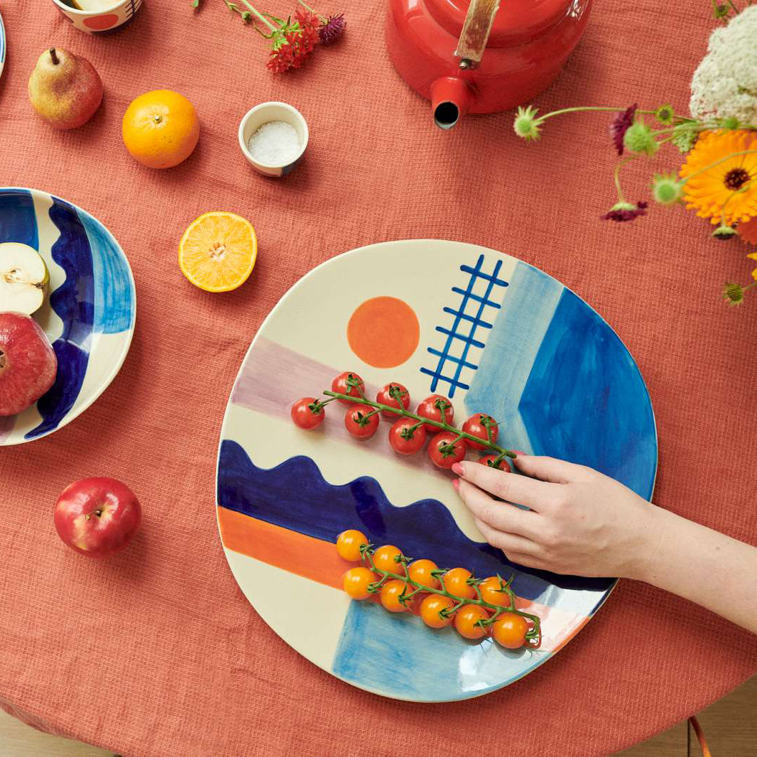 A hand places cherry tomatoes on a large ceramic plate with a colorful landscape design featuring blue waves, an orange sun, and a blue grid. The scene is styled on a textured terracotta tablecloth with other fruits, a red teapot, and another patterned plate.
