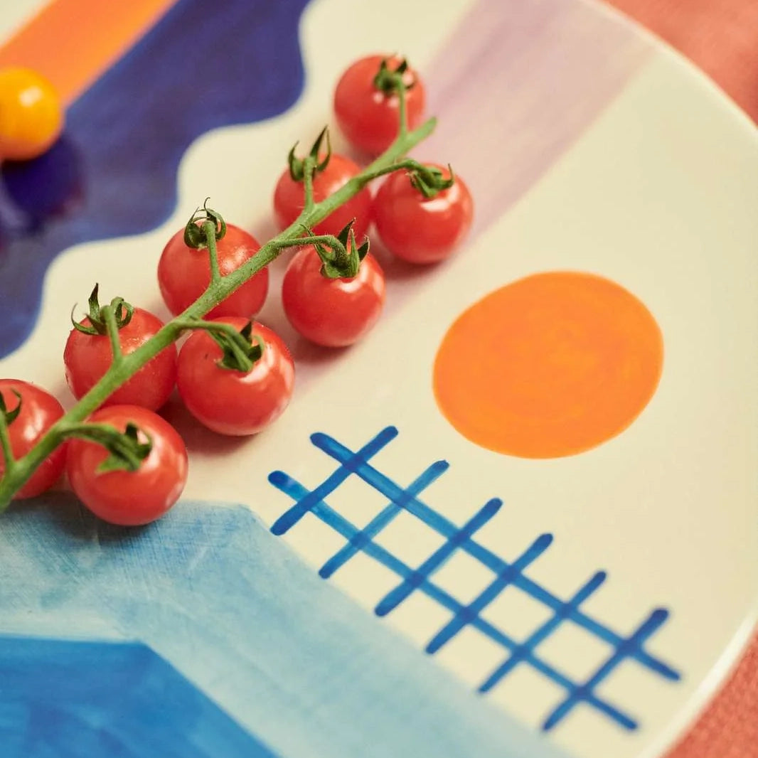 A close-up of a white ceramic plate with a colorful abstract landscape design, featuring a blue grid, blue water, a purple sky, and an orange sun. A vine of red cherry tomatoes is placed on the plate.