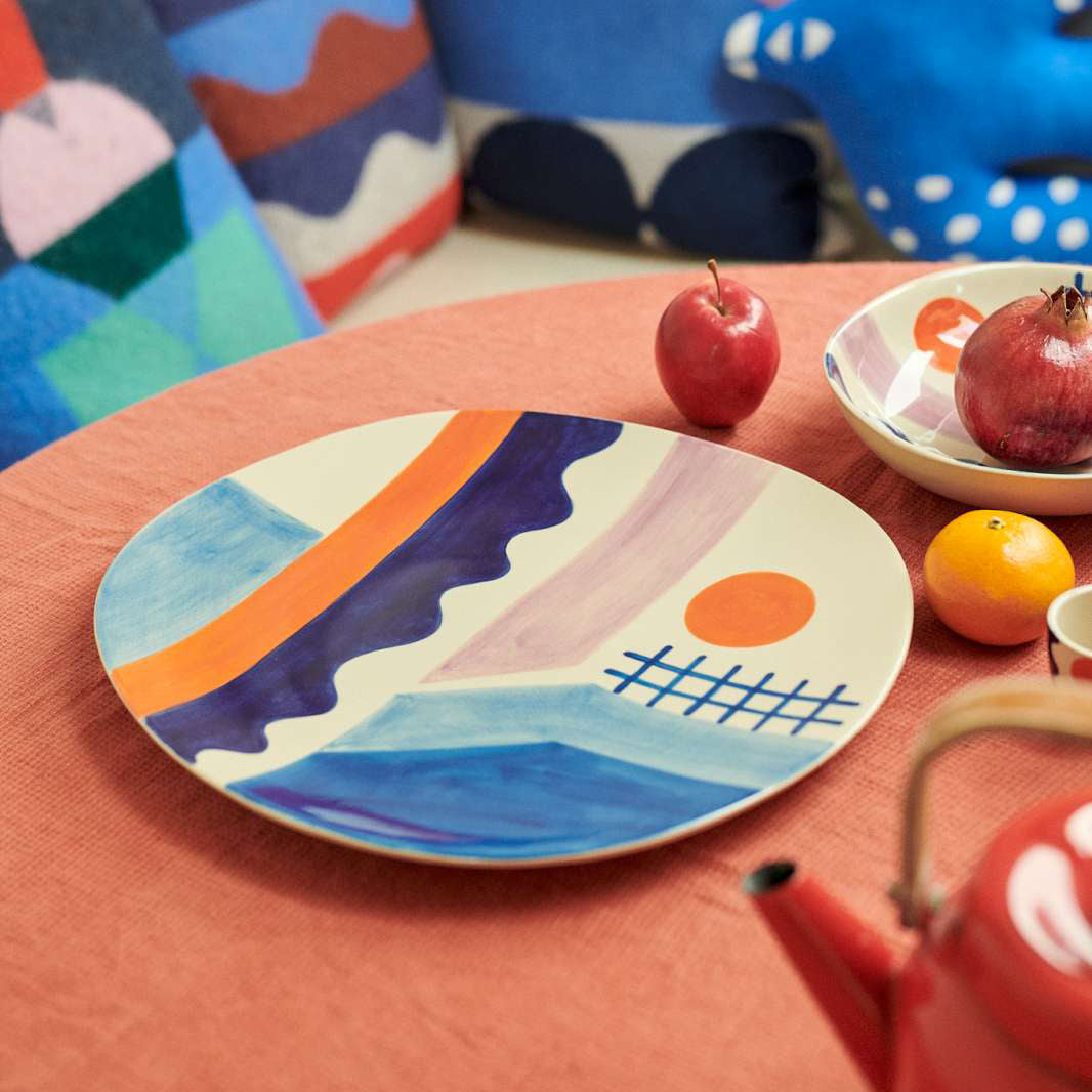 A round ceramic plate with an abstract landscape design in blue, purple, orange, and pink, styled on a terracotta table alongside fruit and colorful cushions in the background.