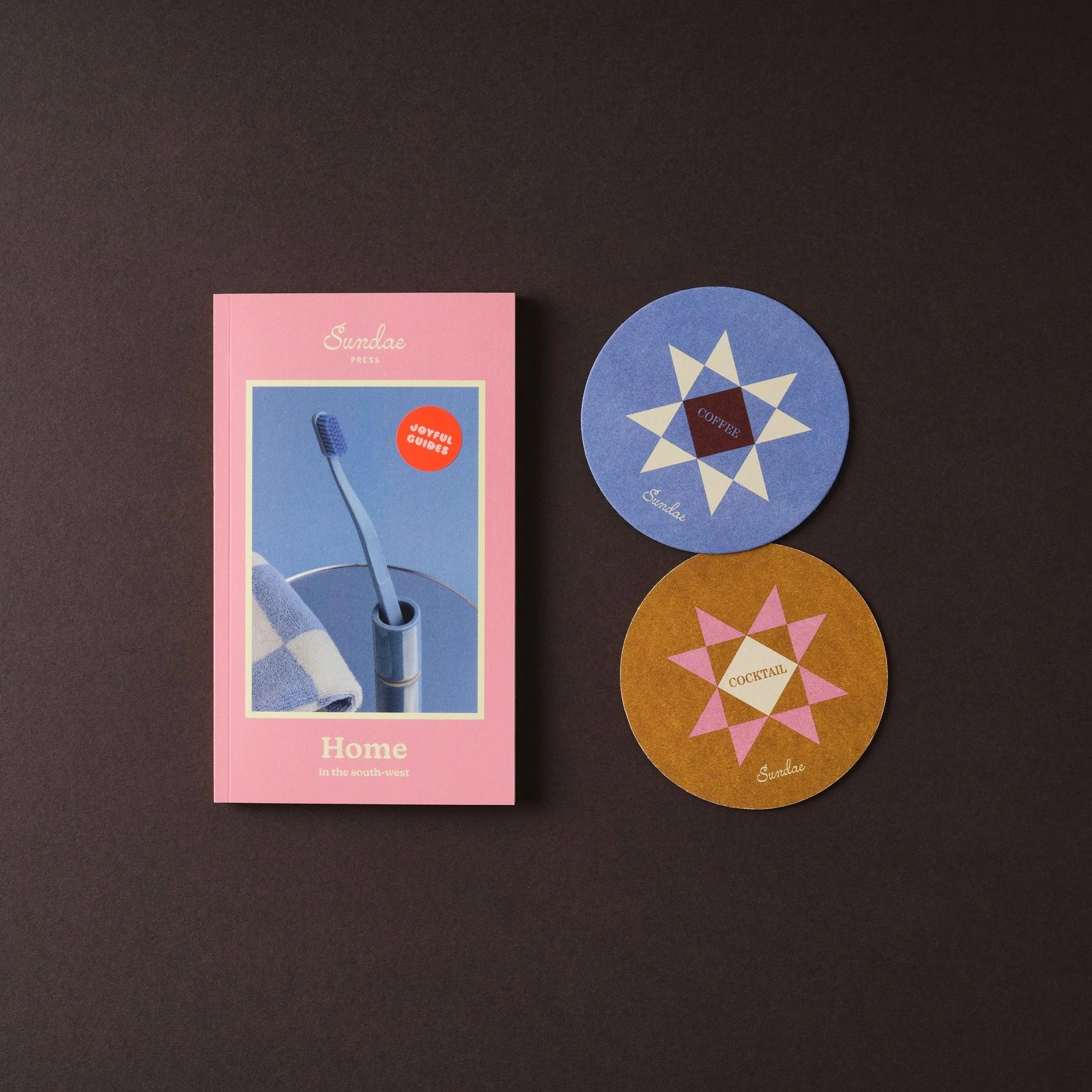 Pink Home guide by Sundae Press with a blue toothbrush on the cover illustration next to two colorful coasters on a dark background
