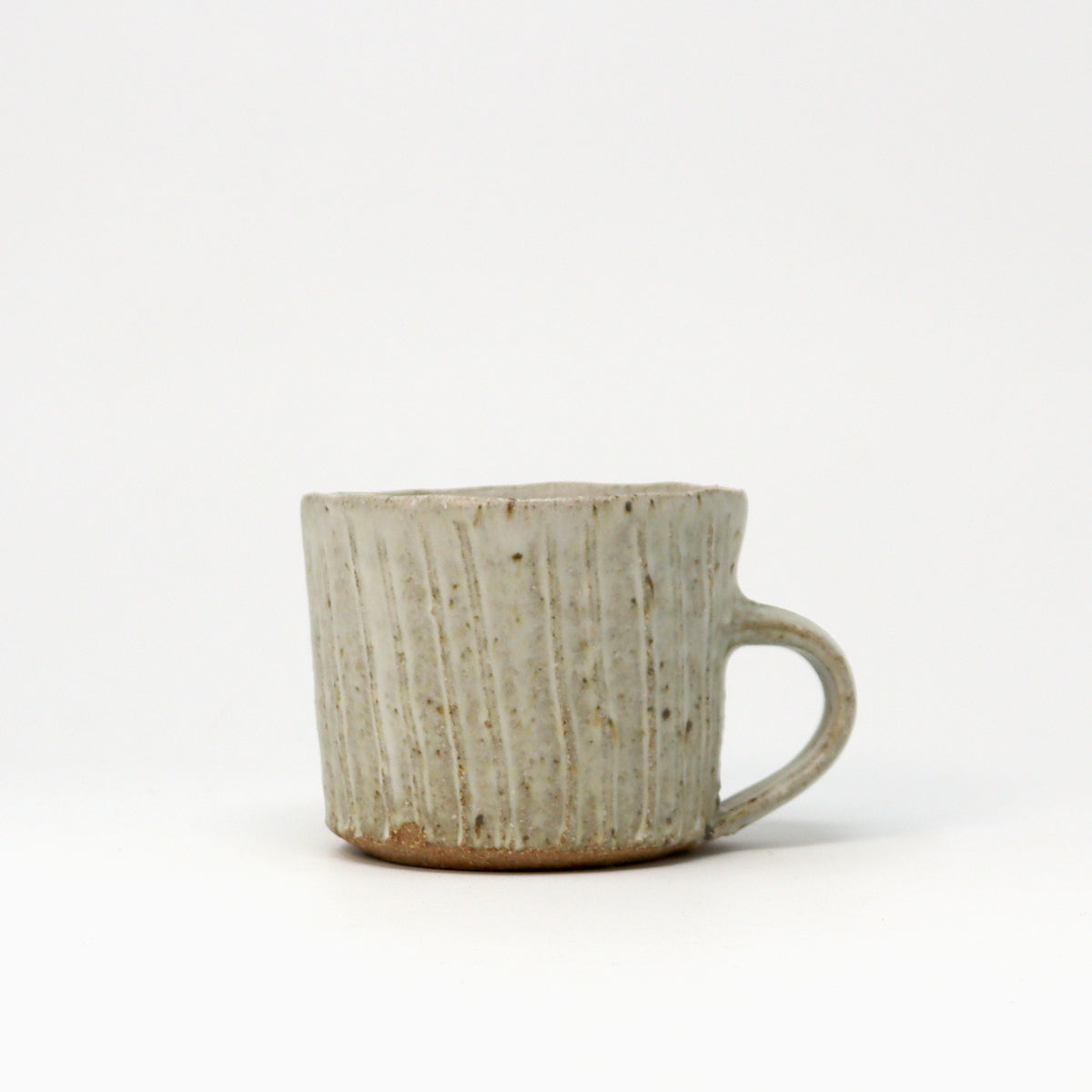 A handmade ceramic cup with a ribbed vertical texture and a C-shaped handle, featuring a speckled light neutral glaze with a natural brown clay base, set against a plain white background.