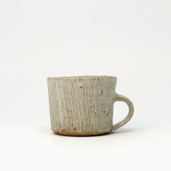 A handmade ceramic cup with a ribbed vertical texture and a C-shaped handle, featuring a speckled light neutral glaze with a natural brown clay base, set against a plain white background.