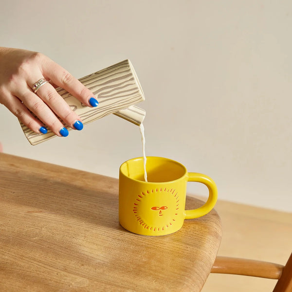 A brown and beige jug in the shape of a log and branch spout with a tree bark design pouring into a yellow sunshine mug on a kitchen table