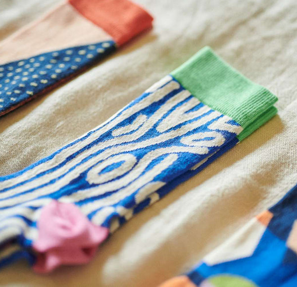 A close-up of a folded knit sock with a blue and white wavy wood pattern, a light green cuff, and a pink fabric accent, laid on a beige textured surface.