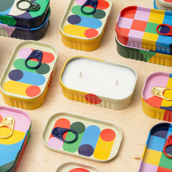 Colorful tin containers with geometric patterns on, containing a candle inside with two wicks on a wooden surface